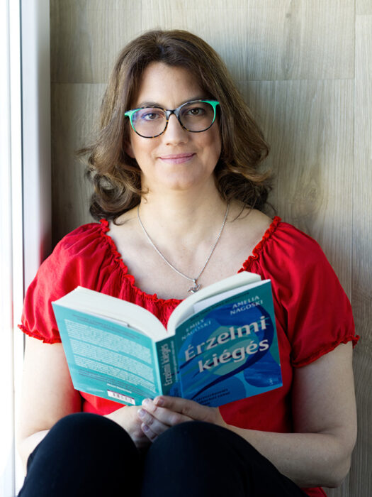 Ez egy fotó: Merényi Katalin Gabriella life és business coach piros rövid ujjú felsőben és zöld-barna keretes szemüvegben felhúzott lábakkal fekete nadrágban egy kék színű könyvet tart nyitva a kezében, miközben ránk mosolyog. A könyv címe: Érzelmi kiégés. A coach nyakában egy ezüst nyakláncon kolibri medál van. A hölgy az ablakpárkányon ül és a falnak támaszkodik.