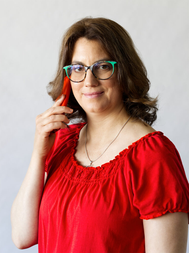 Ez egy fotó: Merényi Katalin Gabriella life és business coach piros érdekes rövid ujjú felsőben, zöld-barna keretes szemüvegben egy piros tokban lévő mobiltelefonon telefonál. A coach a kamerába néz, mosolyog. Haja hullámos, barna és vállig ér. Nyakában egy ezüst kolibri medál, mint a gondolat felszabadulásának szimbóluma.