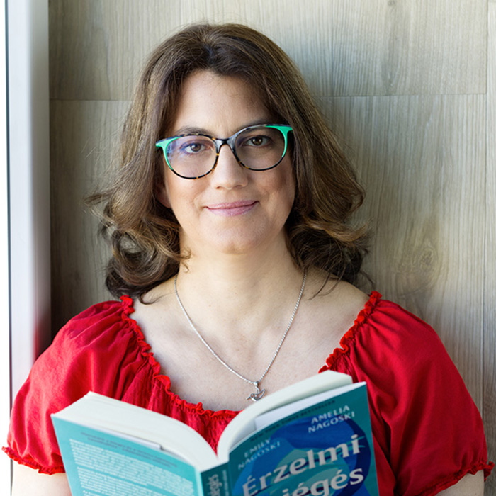 Ez egy fotó: Merényi Katalin Gabriella life és business coach piros rövid ujjú felsőben és zöld-barna keretes szemüvegben egy kék színű könyvet tart nyitva a kezében, miközben ránk mosolyog. A könyv címe: Érzelmi kiégés. A coach nyakában egy ezüst nyakláncon kolibri medál van. A hölgy fotója ívesen van körülvágva, szélei érdekesen egybeolvadnak a háttérrel.